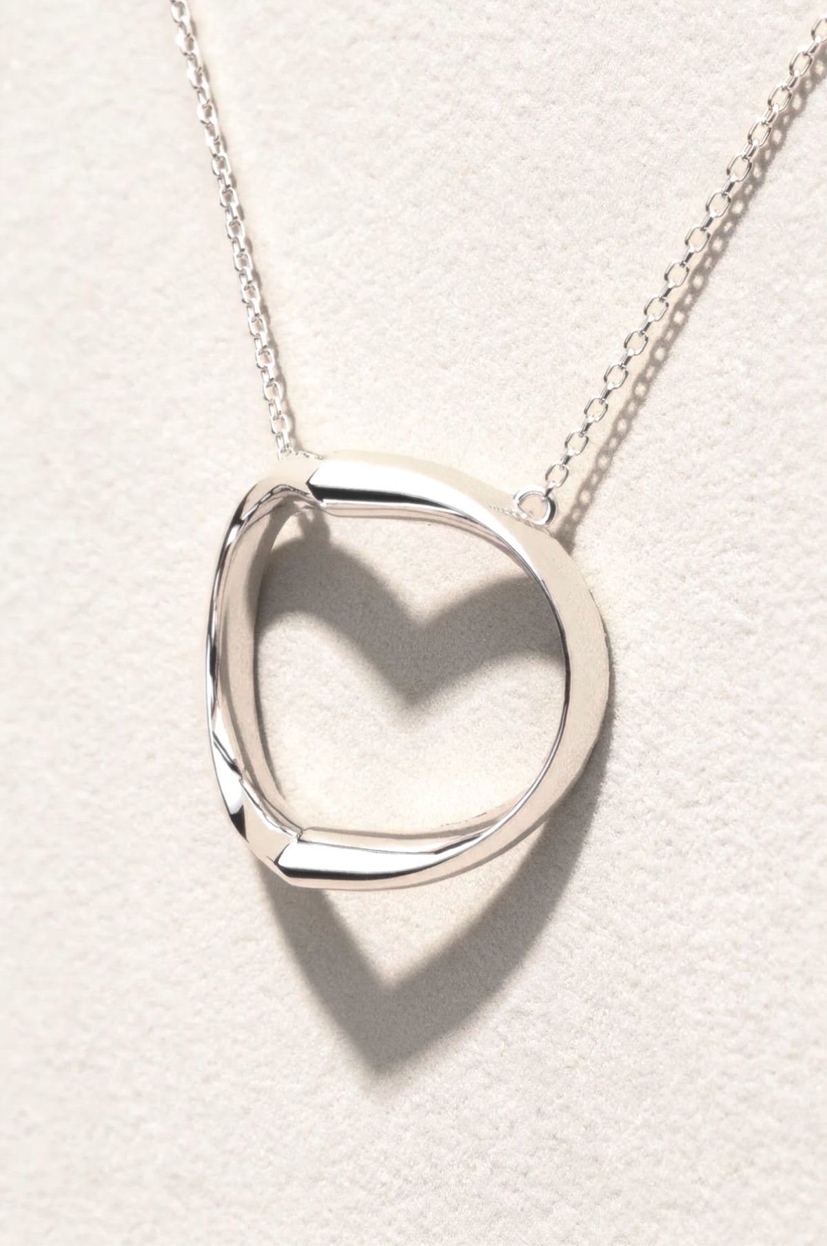 A polished sterling silver pendant necklace suspended on a delicate chain. The sculptural design casts a distinct heart-shaped shadow against a textured off-white background.