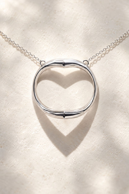 A polished sterling silver necklace with a ring-shaped open pendant that casts a heart-shaped shadow, resting on a textured beige surface with soft lighting.